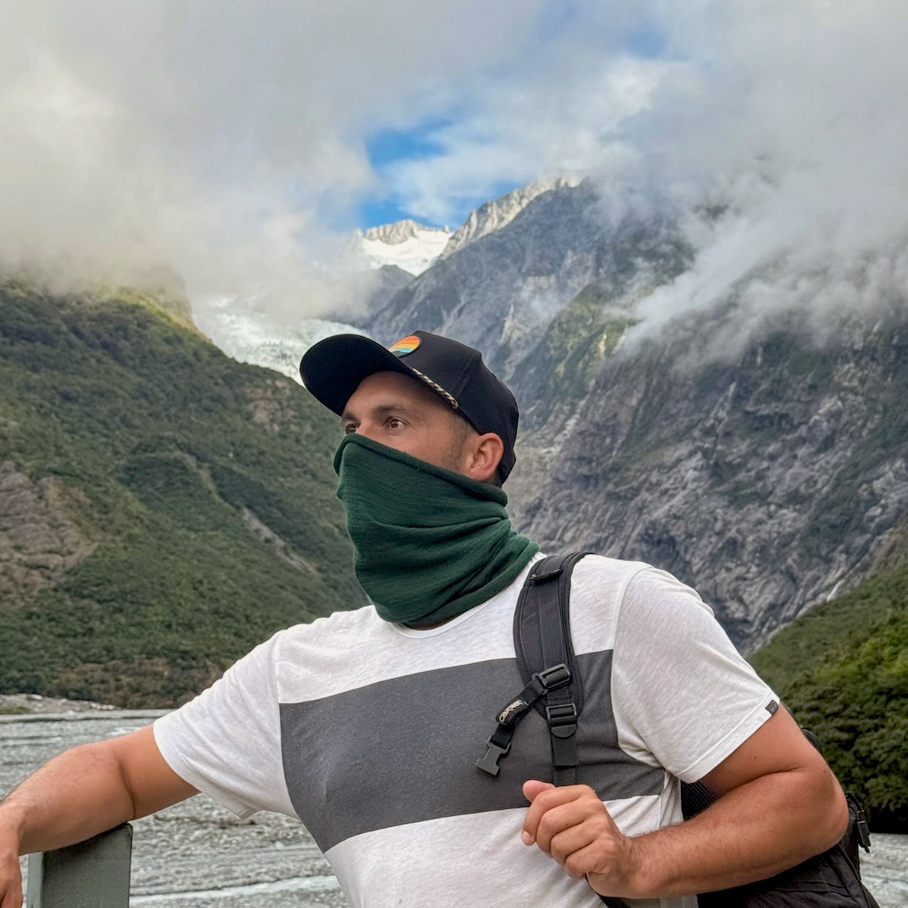 Man with a green face covering and backpack standing in front of a mountainous landscape.