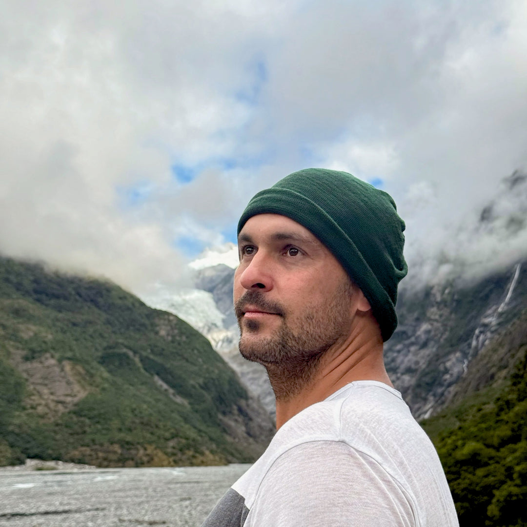 Man wearing a green beanie and white shirt standing in front of a mountainous landscape with clouds.