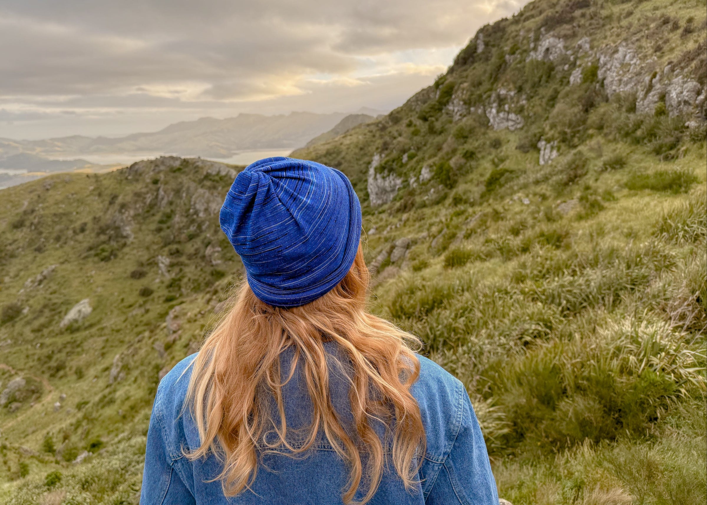 Person wearing a blue beanie and jacket standing on a mountain with a scenic view.