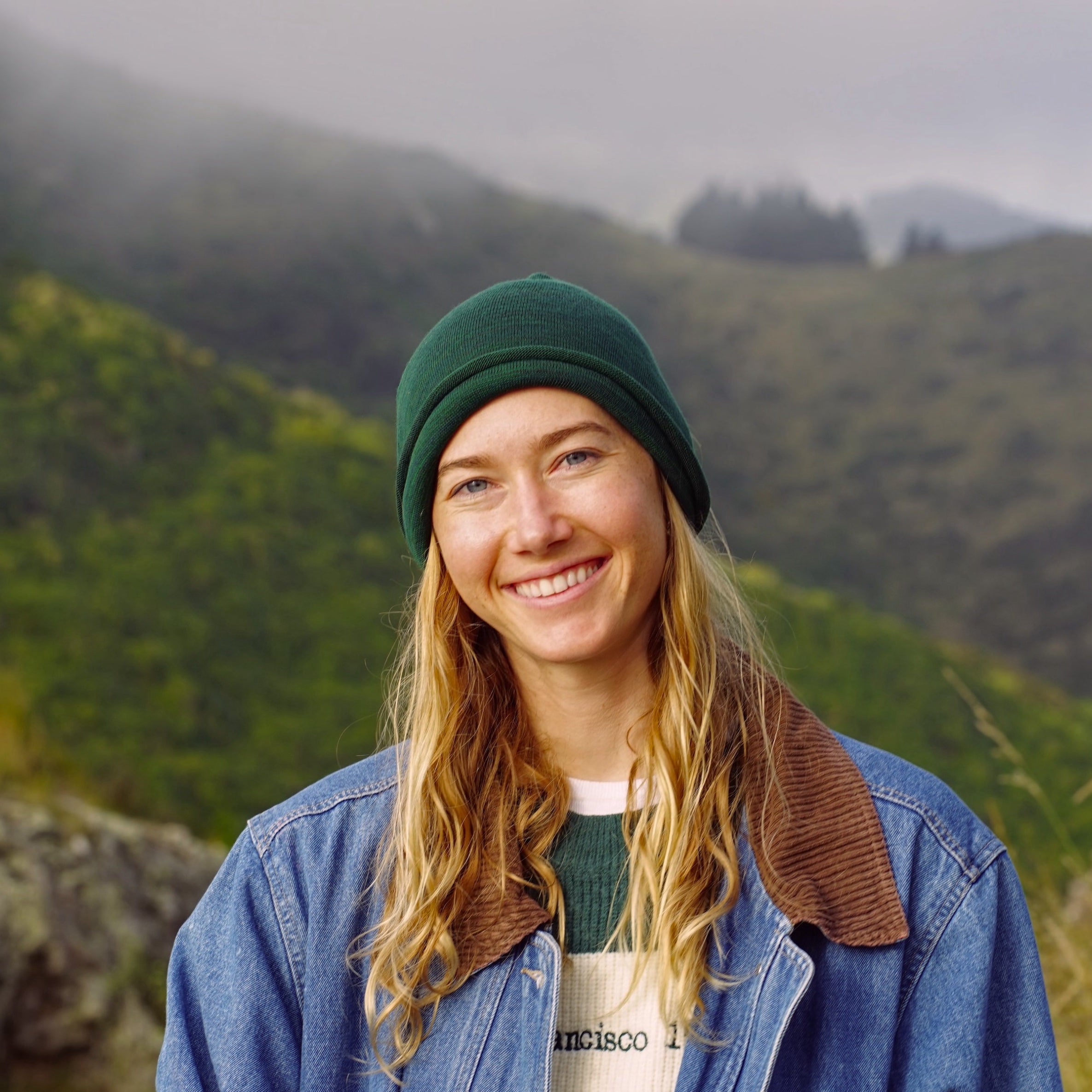 Person wearing a green beanie and blue jacket with a mountainous background